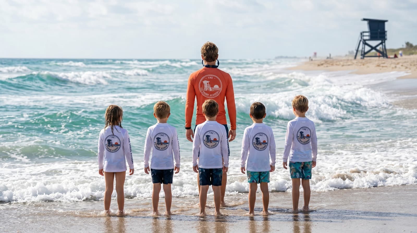 Shore Academy instructor and five students in branded rash guards standing at the ocean's edge at South Florida beach, lifeguard tower visible