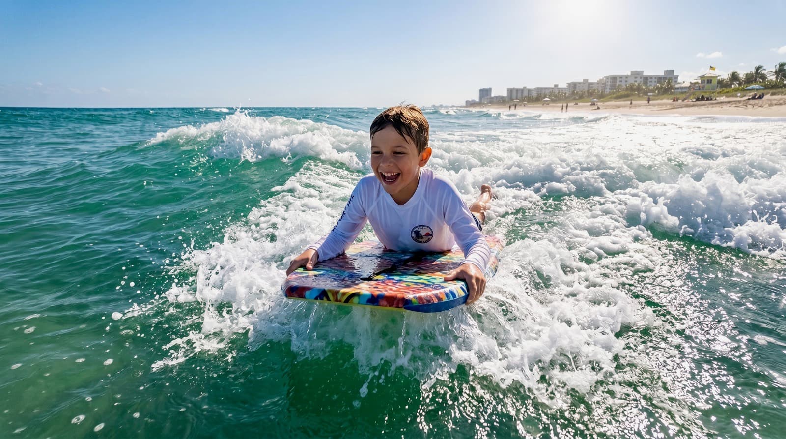 Young boy in Shore Academy rash guard riding a bodyboard through a breaking wave on a sunny South Florida beach, wave navigation training